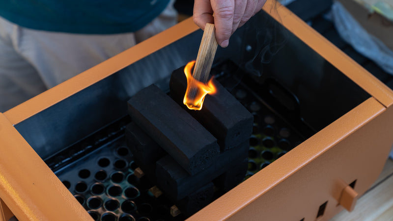 A lit stick of fatwood dropped into a crisscross stack of Thai-Style Charcoal in the Thaan Grill