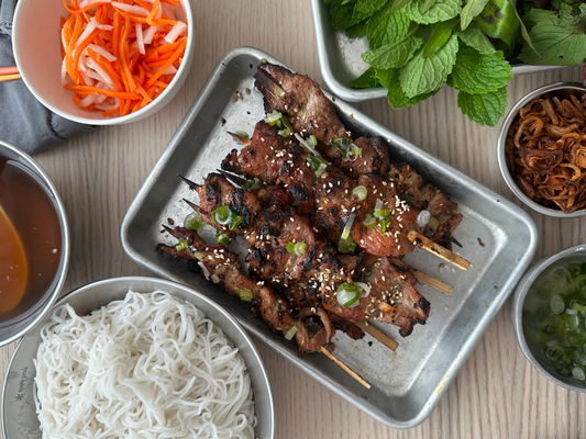 Grilled lemongrass pork shoulder skewers with charred caramelized edges on a sheet pan, surrounded by rice vermicelli, pickled carrots and daikon, fresh herbs, fried shallots, and dipping sauce.