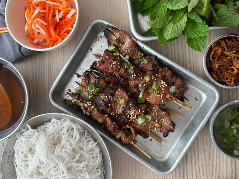 Grilled lemongrass pork shoulder skewers with charred caramelized edges on a sheet pan, surrounded by rice vermicelli, pickled carrots and daikon, fresh herbs, fried shallots, and dipping sauce.