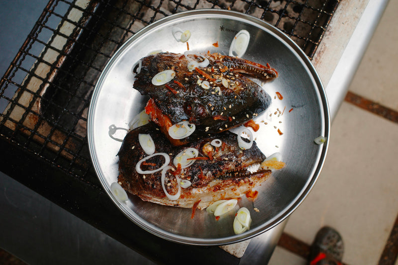 Split salmon head halves glazed with miso-maple butter on a charcoal grill, topped with scallions and sesame seeds